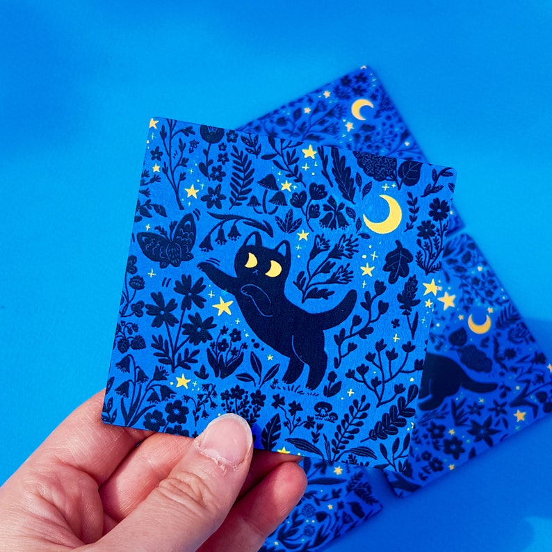 May include: A blue and black illustrated card featuring a black cat with yellow eyes and a crescent moon. The cat is surrounded by a variety of flowers and plants, all in a dark blue color. The card has a whimsical and magical feel.