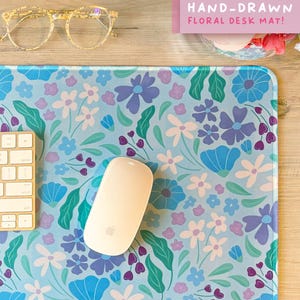 May include: A hand-drawn floral desk mat with a light blue background and a pattern of blue, purple, and white flowers. A white computer mouse and a portion of a keyboard are on the mat. The text "HAND-DRAWN FLORAL DESK MAT!" is visible.