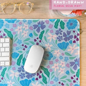 May include: A rectangular desk mat featuring a light blue background with a repeating pattern of flowers and leaves in shades of blue, purple, pink, and green. A pink rectangular label with the text HAND-DRAWN FLORAL DESK MAT! is visible.