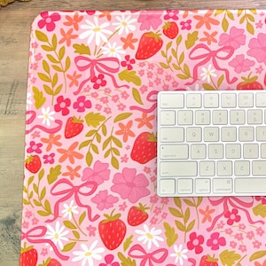 May include: A rectangular desk mat featuring a repeating pattern of red strawberries, pink bows, white daisies, and small pink and orange flowers on a light pink background, with a white computer keyboard placed on the right side of the mat.