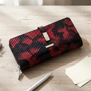 May include: A rectangular wallet featuring a red and black snakeskin-textured pattern. The wallet has a silver-toned metal clasp on a central strap and a silver-toned zipper pull. A small rectangular metal plate with text is centred on the front.