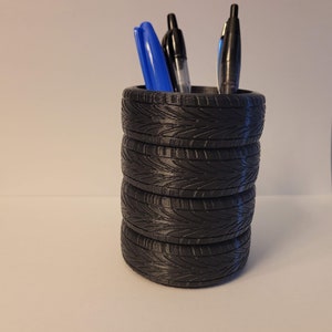 May include: A black tire-shaped pen holder with a stack of tires. Three pens are in the holder, two black and one blue.