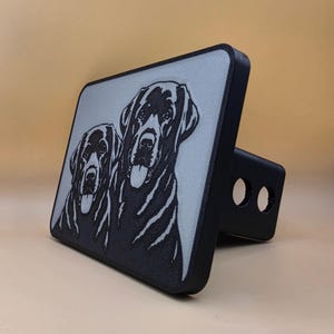 May include: A black and light gray trailer hitch cover featuring a raised graphic of two dogs side by side. The rectangular face displays the detailed black silhouettes of the dogs against a textured light gray background with a black border.