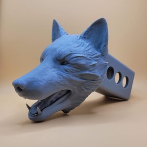 May include: A 3D printed gray wolf head hitch cover. The wolf's mouth is open, showing its teeth. The hitch cover is attached to a gray metal hitch receiver.