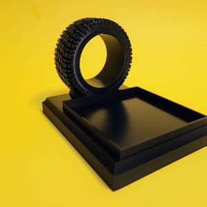 May include: A black phone stand with a tire-shaped holder. The stand has a square base and a raised platform for the phone.
