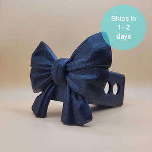 May include: A black bow-shaped trailer hitch cover. The bow is detailed with textured fabric-like patterns. The hitch cover has two circular cutouts. The background is a solid tan color.