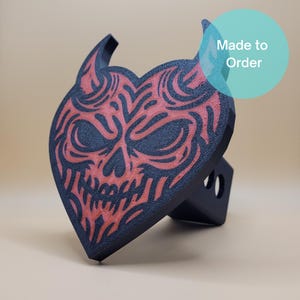 May include: Black and red heart-shaped trailer hitch cover with a devil skull design. The hitch cover has a textured surface and is made to order.