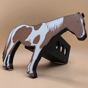 May include: A brown and white horse-shaped decorative item with a black base. The horse has a textured appearance and is supported by a black stand with three circular cutouts. The horse's design features a brown head and body with white patches.