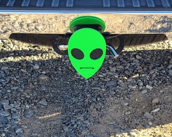 May include: Green alien face hitch cover for a vehicle. The alien has black eyes and a neutral expression.