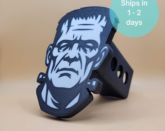 May include: Black and white Frankenstein hitch cover. The hitch cover features a detailed graphic of Frankenstein's face. The product is made of a durable material and is designed to fit standard trailer hitches.