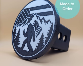 May include: Black and white hitch cover with a Bigfoot design holding an American flag, set against a mountain and tree backdrop. The round cover has the text "Made to Order".