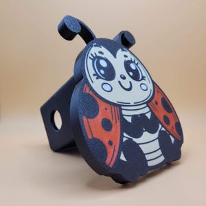 May include: A black and red ladybug hitch cover with a cute cartoon face and big eyes. The ladybug is smiling and has black spots on its wings.