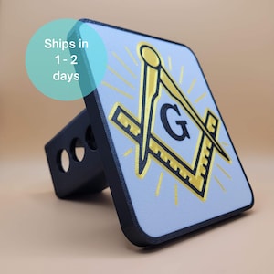 May include: A black and gold Masonic Square and Compass hitch cover. The hitch cover features a light blue background with a gold Square and Compass symbol and the letter "G". The hitch cover is rectangular with a black border.