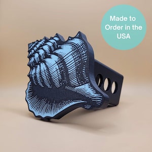 May include: A black trailer hitch cover featuring a detailed light blue seashell design. The hitch cover has three holes and the text "Made to Order in the USA" on a teal circle.