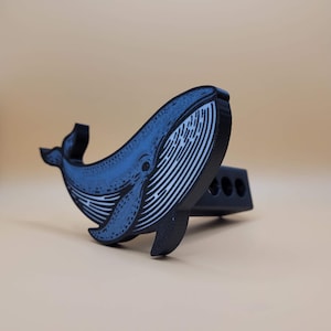 May include: A black and blue whale-shaped trailer hitch cover. The whale has a detailed design with white stripes and a blue body. The hitch cover is designed to fit into a standard 2-inch receiver.