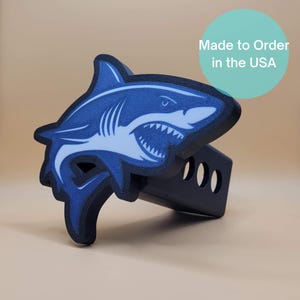 May include: A blue and white shark design on a black trailer hitch cover. The shark graphic features sharp teeth and is set against a light background. The text "Made to Order in the USA" is in a teal circle.