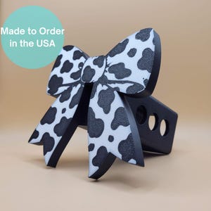 May include: A black and white cow print bow-shaped trailer hitch cover. The bow has a textured surface and is made of a durable material. The hitch cover has three circular holes. Text in a teal circle reads "Made to Order in the USA."