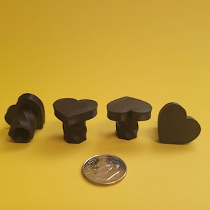 May include: Four black heart-shaped table legs with a spiral design. The table legs are arranged in a row on a yellow surface. A US dime is in the foreground for scale.