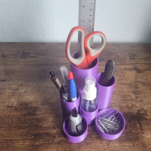 May include: Purple desk organizer with six compartments. The organizer holds a pair of red scissors, a ruler, a glue stick, a bottle of hand sanitizer, a few pens, and a container of paper clips.