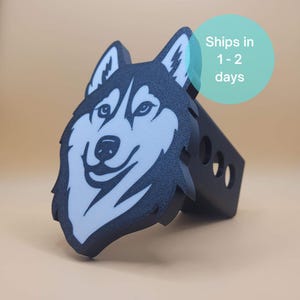 May include: A black and white husky dog head design on a trailer hitch cover. The hitch cover has a textured surface and a black base with three circular holes. The text "Ships in 1-2 days" is in a blue circle.