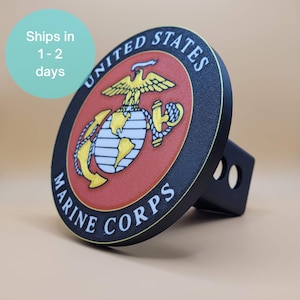 May include: Black trailer hitch cover featuring the United States Marine Corps emblem. The emblem is a circular design with a red background, the words "United States Marine Corps", and a gold eagle, globe, and anchor.