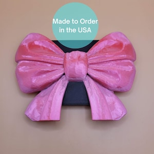 May include: A large, pink bow with a textured surface, mounted on a black rectangular base. The bow has a gradient of pink hues, from light to darker shades. Text in a blue circle reads "Made to Order in the USA."