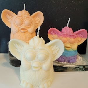May include: Three handmade candles in the shape of a Furby toy. One is ivory, another is peach, and the third is a rainbow of colours. Each candle has a wick and is placed on a white surface.