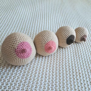 May include: Four handmade crochet breast-shaped pillows in a row. The pillows are beige with pink or brown areolas. The pillows are arranged on a white, textured surface. The pillows vary slightly in size and color.