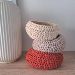 Puede incluir: Una pila de tres cestas de ganchillo en colores neutros. La cesta superior es beige, la del medio es malva claro y la inferior es naranja quemado. Las cestas están apiladas una encima de otra.