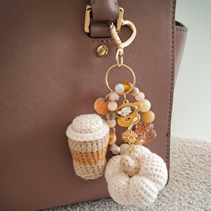 Op de afbeelding: Een goudkleurige metalen sleutelhanger met een ronde ring en diverse kralen in beige- en oranjetinten. Het bevat een kleine oranje pompoenbedel, een goudkleurige koffiekopbedel, een gehaakte koffiebeker en een witte gehaakte pompoen.