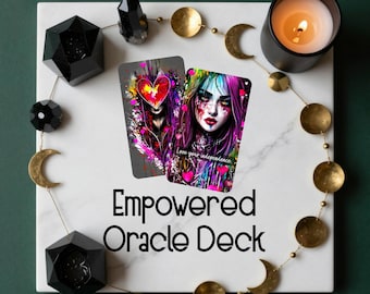 May include: An "Empowered Oracle Deck" is displayed on a white marble surface, accompanied by a lit candle in a black holder and decorative gold-colored moons and circles. Two oracle cards feature colorful illustrations of a woman and a heart.