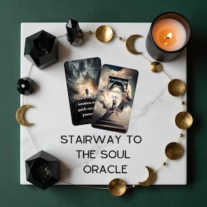 May include: Two oracle cards titled Stairway to the Soul Oracle are displayed on a white marble surface. The cards feature a person walking up stairs toward a bright light. A black candle, black geometric shapes, and a gold moon phase garland surround them.