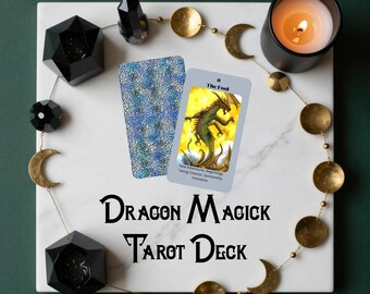 May include: A Dragon Magick Tarot Deck is displayed on a white marble surface, surrounded by decorative elements. The deck includes a card featuring a dragon illustration and the text "The Fool." A lit candle and other mystical accents complete the scene.