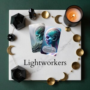 May include: Two rectangular oracle cards featuring blue and green alien-like figures, surrounded by a gold moon phase garland, two black geometric candle holders, and a lit candle on a white marble board with the text Lightworkers printed below.