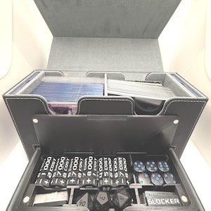 May include: A gray, multi-compartment game box with a hinged lid. The box contains card decks, dice, and cost counters. The cost counters are black with white text. The dice are black with blue dots. The box is made of a textured material.