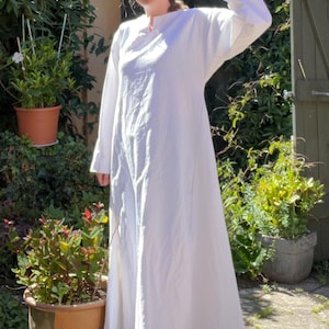 Puede incluir: Un vestido blanco largo hasta los pies con mangas largas y un escote redondo sencillo. La prenda presenta una silueta holgada y fluida que llega hasta los tobillos, confeccionada en una tela blanca lisa con una textura suave y sin adornos.