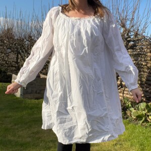 Può includere: Un abito bianco a maniche lunghe con scollatura arricciata e silhouette ampia e fluida. L'abito arriva al ginocchio e viene indossato con leggings neri. L'immagine è scattata all'aperto in una giornata di sole, con erba verde e cielo blu sullo sfondo.