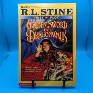 May include: A paperback book titled "Golden Sword of Dragonwalk" by R.L. Stine. The cover features a person with outstretched hand, with the title in gold lettering. The background is a dark forest scene with swirling yellow and green accents.