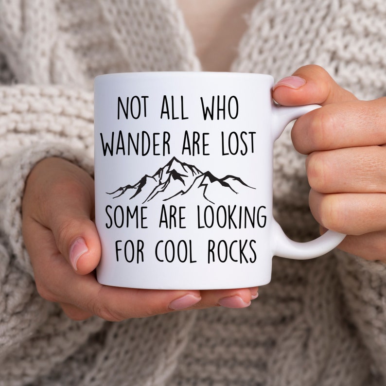 May include: White ceramic mug with a black mountain range graphic and the text "Not all who wander are lost, some are looking for cool rocks".