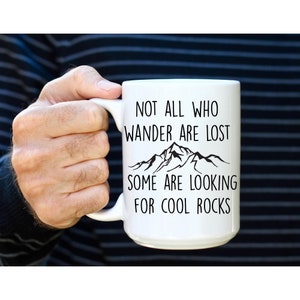 May include: White ceramic mug with a black mountain range graphic and the text "Not all who wander are lost, some are looking for cool rocks."
