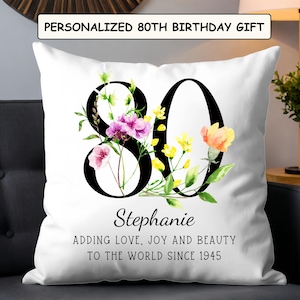 May include: White throw pillow with a black "80" design, adorned with watercolor flowers in pink, yellow, and orange. The text "PERSONALIZED 80TH BIRTHDAY GIFT" is at the top. Below the "80" is the name "Stephanie" and the phrase "ADDING LOVE, JOY AND BEAUTY TO THE WORLD SINCE 1945."