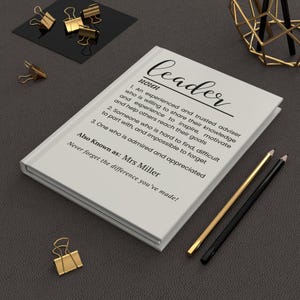 May include: A white journal with the definition of "leader" printed on the cover. The definition includes the text "Also Known as: Mrs Miller" and "Never forget the difference you've made!" Gold binder clips and pencils are arranged around the journal.