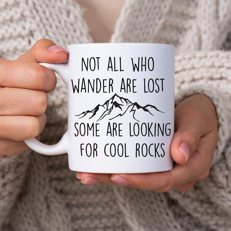 May include: White ceramic mug with black text and a mountain range graphic. The text reads "Not all who wander are lost, some are looking for cool rocks."