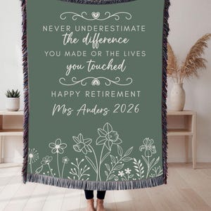 May include: A sage green woven throw blanket featuring white text that reads: NEVER UNDERESTIMATE the difference YOU MADE OR THE LIVES you touched HAPPY RETIREMENT Mrs Anders 2026. The blanket has a fringed edge and a white floral line art design.