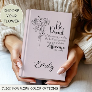 May include: A pale pink journal featuring a floral illustration and the words "Be Proud" and "Emily" in black script. The cover also includes the text "of the work you do, the brilliant person you are and the difference you make."