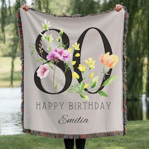 May include: A light gray woven throw blanket with the number 80 in black, decorated with watercolor flowers in pink, yellow, and orange. The words "HAPPY BIRTHDAY Emilia" are printed below the number. The blanket has a fringed edge.