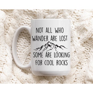 May include: White ceramic mug with a black graphic that reads "Not all who wander are lost, some are looking for cool rocks". The graphic also includes a stylized image of mountains.