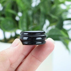 May include: A small, black, round object with a tiered design is held in a person's hand. The object has a glossy finish and appears to be made of a solid material. The background is blurred, featuring green foliage.