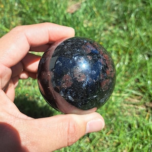May include: A polished black sphere with red and silver flecks. The sphere is made of a natural stone and is approximately 2 inches in diameter.