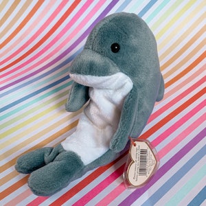 May include: A gray and white plush dolphin toy with a white belly and a gray back. The toy has a Ty heart-shaped tag attached to it that says "Please remove all swing tags before giving this item to a child."